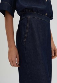Dark blue denim pants with a smooth texture, featuring contrasting yellow stitching, a side pocket, and a wide waistline.