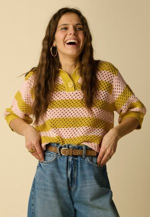 Jonge vrouw met lang golvend haar lacht, draagt grote oorringen, een roze en geel gestreepte gebreide trui, hoge taille jeans en een bruine riem.