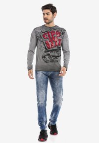 Graues Langarmshirt mit auffälligem rotem und schwarzem Grafikdruck. Trägt dazu eine blaue Jeans und schwarze Sneakers mit roten Akzenten.
