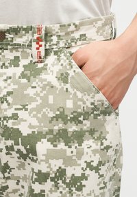 Χέρι που ακουμπάει μέσα στην τσέπη παντελονιού ψηφιακής παραλλαγής σε πράσινο και μπεζ, με λεπτομέρειες θηλιάς ζώνης κόκκινου και άσπρου χρώματος.