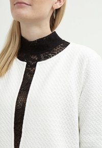 Vit texturerad jacka med rund halsringning över en svart spets-topp med hög krage, som har intrikata mönster och en slät yta.