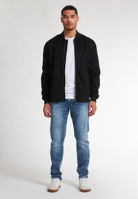 Jachetă bomber din suplex negru cu fermoar frontal, asociată cu blugi albastre deschise și adidași albi. Modelul stă în fața unui fundal neutru.