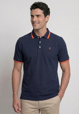 Hombre que lleva una camiseta polo azul marino con rayas naranjas y rojas en el cuello y las mangas, sonriente, pantalones beige, mano en el bolsillo.