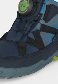 Gros plan d’une chaussure de sport bleu foncé avec système de laçage Boa et des brides en tissu noir et bleu sur les orteils et le dessus du pied.