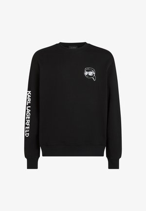Musthave musta T-särk, millel on valge "Karl Lagerfeld" tekst paremal varrukal ja väike joonistatud nägu, mis kannab prille, vasakul rinnal.