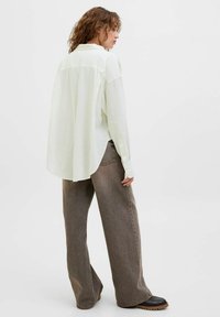 Camicia oversized verde chiaro in tessuto morbido, con colletto a bottoni, maniche lunghe e orlo arrotondato; abbinata a jeans marroni larghi.