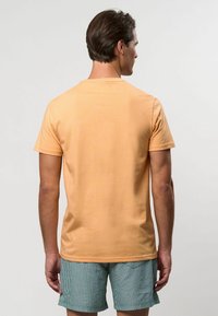 Īsām piedurknēm, gaiši oranža kokvilnas T-kRekle ar apaļu kakla izgriezumu; skatīta no aizmugures, kopā ar zaļām svītrainām šortiem.