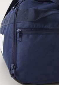 Sac de sport bleu marine fabriqué dans un tissu texturé, doté d'une poche zippée et d'une poignée résistante. Design minimaliste sans logos visibles.