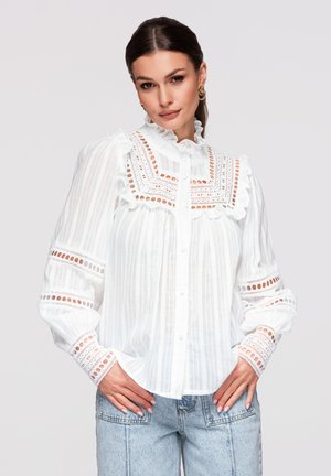 Donna con capelli scuri indossa una camicetta bianca in pizzo con colletto e polsini arruffati, abbinata a jeans azzurri a vita alta, mani sui fianchi.