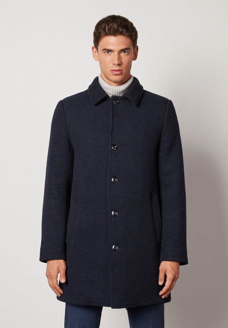 Marinblå ullrock med knäppning framtill, spetsig krage, sidofickor och rak skärning. Slät textur och diskret design.