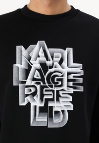 Svart sweatshirt med fet, tredimensionell vit textdesign som stavar "KARL LAGERFELD," med varierande skuggning och texturerad look.