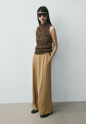 Donna con capelli lunghi e scuri che indossa occhiali da sole oversize, top lavorato a maglia senza maniche, pantaloni beige larghi e scarpe nere, in piedi con le mani in tasca.