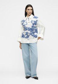 Femme portant un chemisier blanc avec de larges poignets, un gilet tricoté bleu et blanc à motifs floraux, un jean bleu clair à jambes larges et des chaussures noires à bouts pointus avec des nœuds.
