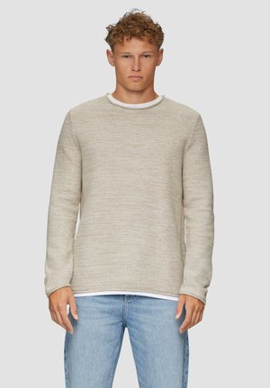 Lichtbeige sweater, textuur gebreide stof, ronde halslijn en lange mouwen, gelaagd over een wit shirt, gecombineerd met lichtblauwe jeans.