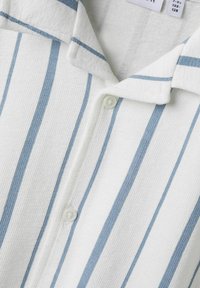 Camicia bianca con strisce blu, caratterizzata da un tessuto strutturato, colletto corto e chiusura a bottoni. Cuciture di alta qualità visibili vicino ai bottoni.