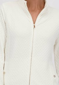 Femme portant une veste zippée blanche texturée avec une fermeture éclair argentée et deux poches à bouton-pression sur les côtés.