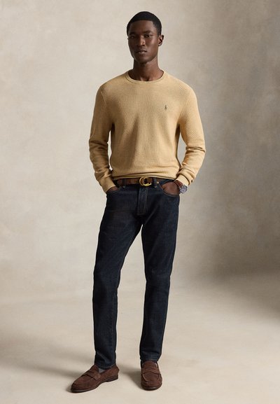 Uomo in piedi con le mani nelle tasche, indossa un maglione beige, jeans scuri, cintura marrone e mocassini, e un orologio da polso su uno sfondo semplice.