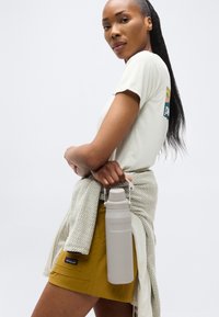 Γυναίκα με μακριά μαλλιά σε casual ντύσιμο κρατώντας ένα ανοιχτό γκρι μπουκάλι νερού από τη λαβή του πίσω από την πλάτη της σε απλό φόντο.