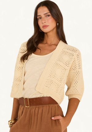 Femme aux longs cheveux bruns porte un cardigan au crochet crème, un haut beige, une ceinture marron et une jupe sable, debout avec une main dans la poche.
