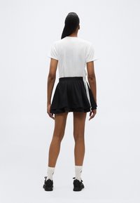 Άτομο που στέκεται με την πλάτη του προς τα εμάς, φορώντας λευκό t-shirt, μαύρη φούστα μίνι με στρώσεις και λευκές ρίγες, λευκές κάλτσες και μαύρα αθλητικά παπούτσια.