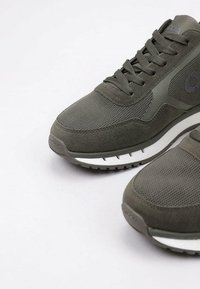 Baskets de sport vert olive avec des matériaux en mesh et en daim, forme arrondie, surface texturée et semelles en caoutchouc blanc avec des rainures.