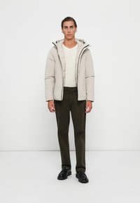Giacca isolante color beige chiaro con cappuccio, chiusura con zip e polsini elastici, indossata sopra una camicia bianca e abbinata a pantaloni verde scuro e scarpe nere.