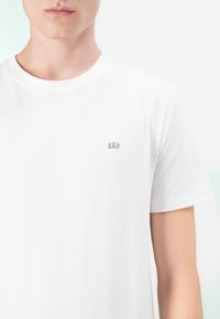 Junge Person trägt ein schlichtes weißes T-Shirt mit kurzen Ärmeln und einem kleinen "GAP"-Logo auf der linken Brust vor einem schlichten weißen Hintergrund.