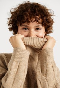 Maglione beige in maglia con colletto alto a coste, caratterizzato da motivi a treccia testurizzati e un tessuto morbido e accogliente, mani che tengono il colletto.