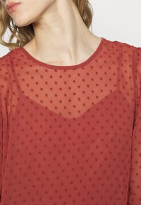 Blusa vermelha transparente com um padrão de bolinhas texturizado, decote redondo e um design em camadas que cria um visual sutil e elegante.