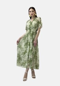 Grünes Blumen-Maxi-Kleid mit kurzen, flatternden Ärmeln, gerafftem Taillenteil und leichtem, durchsichtigem Stoff. Kombiniert mit weißen, schmalen Sandalen.