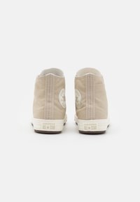 Para beżowych sneakersów typu high-top Converse All Star ukazanych od tyłu na jednolitym białym tle.