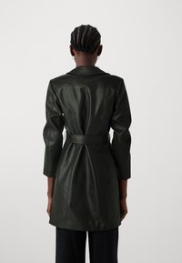 Abrigo de cuero verde oscuro con cintura ceñida, corte entallado y mangas tres cuartos. Textura suave y elementos de diseño estructurados.