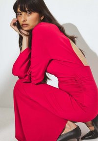 Punainen pitkähiha dressi, jossa on avoin selkä ja kerättyä yksityiskohtaa sivuilla, valmistettu sileästä kankaasta. Mustat korkokengät viimeistelevät asun.