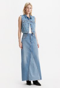 Denimvest met een cropped ontwerp over een witte top, gecombineerd met een lange blauwe denimrok met een diagonale naad en rafelige zoom. Zwarte laarzen.
