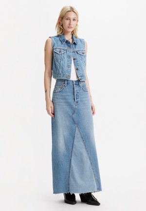 Jeansvest med en cropped design over en hvit topp, kombinert med en lang blå denimskjørt med diagonal søm og frynset kant. svarte støvler.