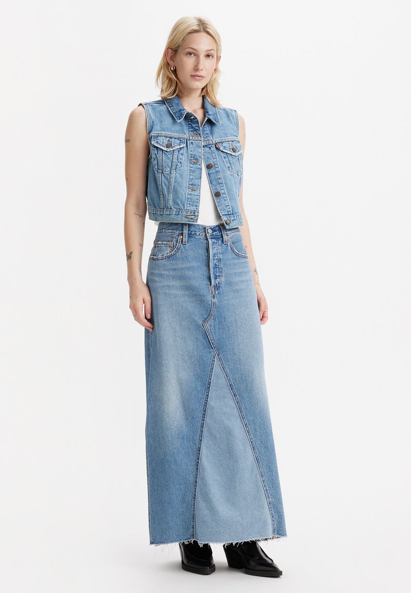 Denimvest met een cropped ontwerp over een witte top, gecombineerd met een lange blauwe denimrok met een diagonale naad en rafelige zoom. Zwarte laarzen.