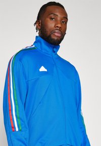 Giacca sportiva blu con colletto alto, caratterizzata da strisce multicolori (rosse, bianche, verdi) lungo le maniche e un logo bianco.