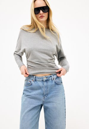 Pikad blond juuksed omav naine, kandes musti päikeseprille, halli pikkade varrukatega pluusi ja siniseid teksapükse, kohendab oma särgi äärt.