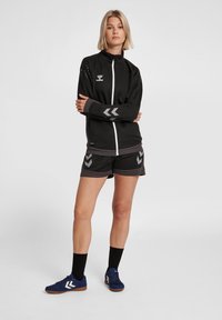 Veste de sport noire avec fermeture éclair blanche, logo argenté et accents en chevron. Associée à un short noir avec un détail en chevron assorti. Chaussures bleu marine.