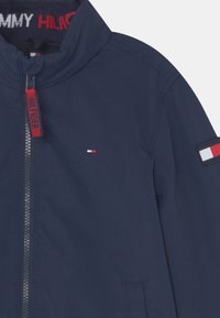 Mörkblå vindjacka med hög krage, hel dragkedja, liten logotyp på bröstet, samt färgblockerade detaljer på ärm och inre krage.