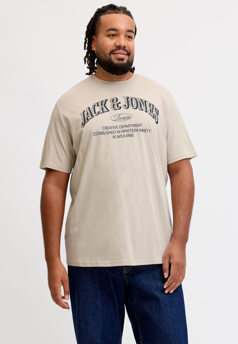 Άνδρας με πλεξούδες που φοράει μπεζ T-shirt Jack & Jones και σκούρο μπλε τζιν, στέκεται μπροστά σε απλό λευκό φόντο.