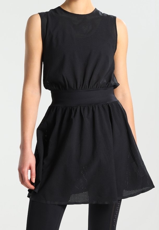 Une femme porte une robe de sport noire sans manches avec une taille froncée et une jupe évasée par-dessus des leggings noirs, se tenant devant un fond neutre.