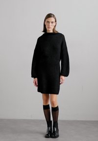 Vestido de malha preto com gola alta, mangas oversized e silhueta reta. Combinado com meias pretas até ao joelho e botas de tornozelo.
