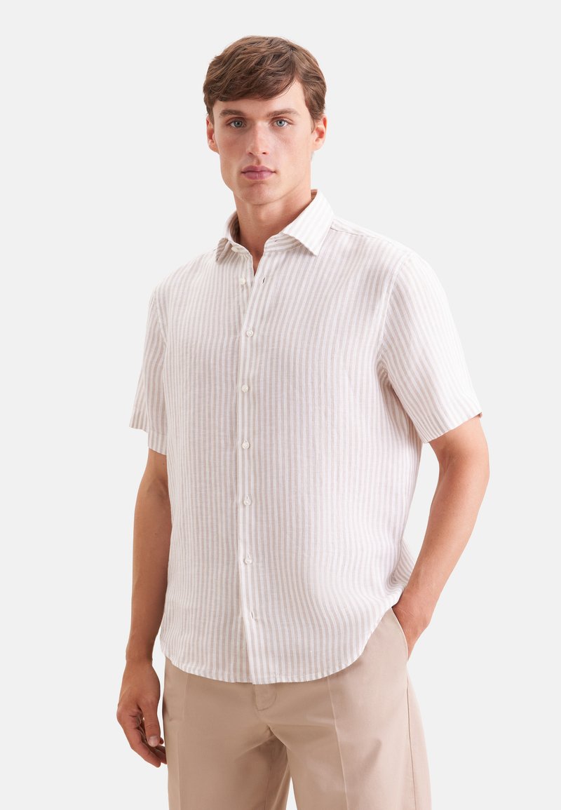 Homme portant une chemise beige rayée à manches courtes et un pantalon beige clair, debout avec une main dans la poche sur fond blanc.