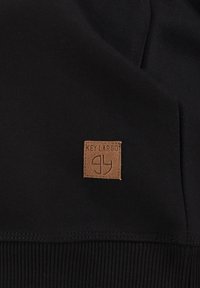Tela negra con textura acanalada y puños. Un parche cuadrado de cuero marrón claro presenta "KEY LARGO" y un símbolo de logo.
