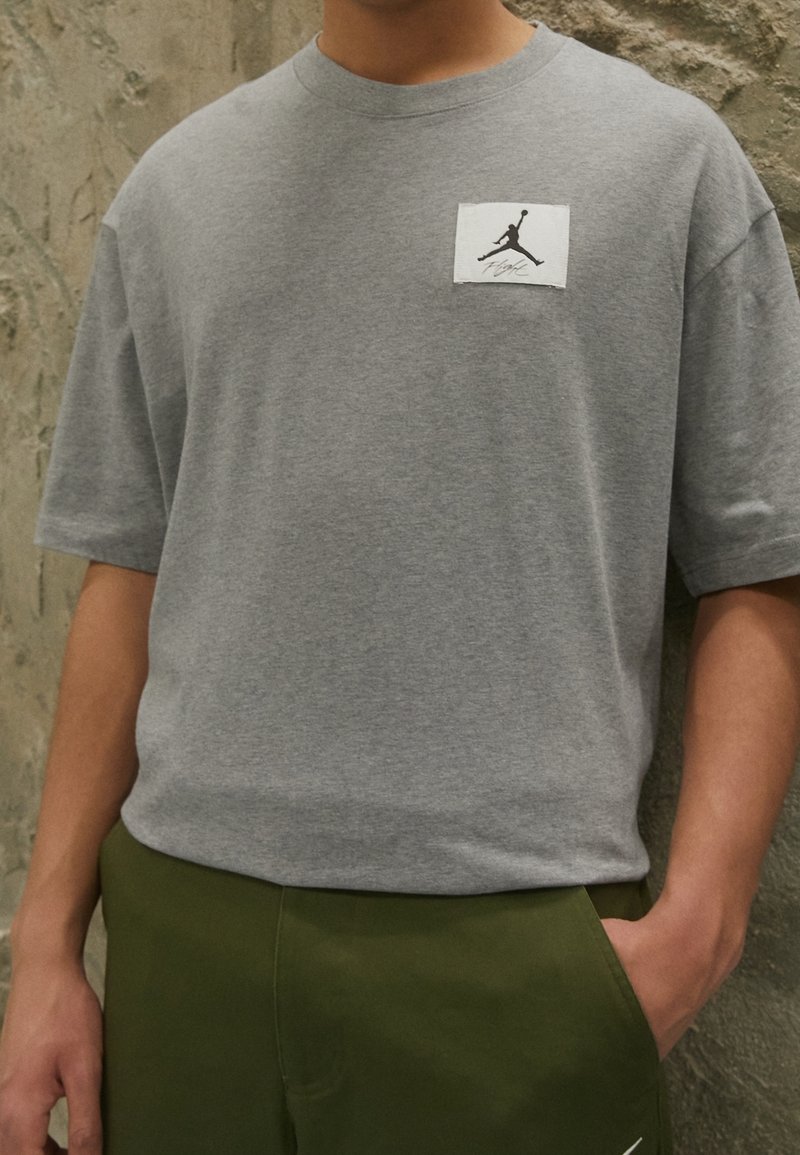 T-shirt gris à manches courtes en mélange de coton, présentant un patch carré blanc avec un logo Jordan noir. Porté avec un short vert olive.
