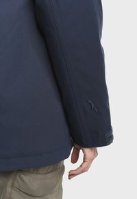 Persona che indossa una giacca blu navy con un piccolo logo ricamato sulla manica e pantaloni beige, con la mano rilassata al lato.