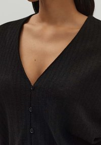 Černý žebrovaný kardigan s hlubokým výstřihem do V a šesti knoflíky. Tkanina se zdá být měkká s mírně strukturovaným povrchem a uvolněným střihem.