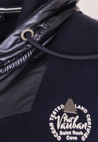 Maglione blu navy con un pannello in nylon nero lucido, coulisse regolabile e logo circolare con la scritta "Port Vauban".
