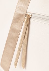 Crème-kleurige synthetische tote bag met een textuur oppervlak, met een gouden lint schouderband en een ritssluiting pocket met een kwastje detail.